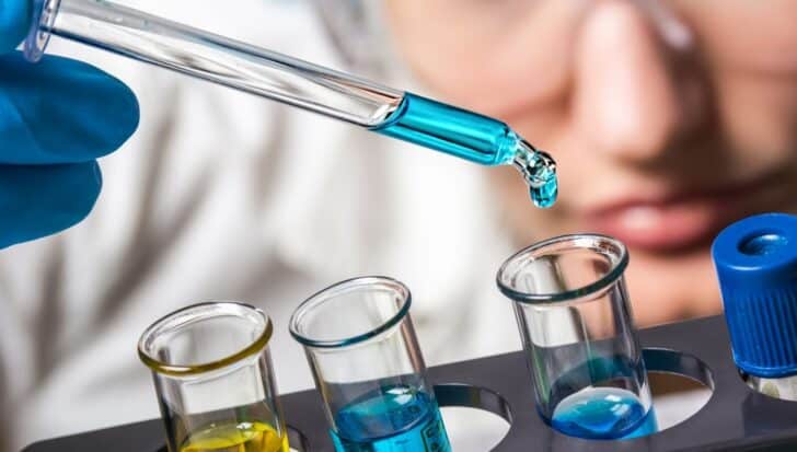 Close-up of a person in safety goggles using a pipette to add blue liquid to test tubes, likely related to significant chemical reactions